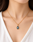 A close-up of a woman wearing a delicate gold chain necklace with a teardrop-shaped pendant featuring a deep blue enamel background and a white tooth motif in the center.