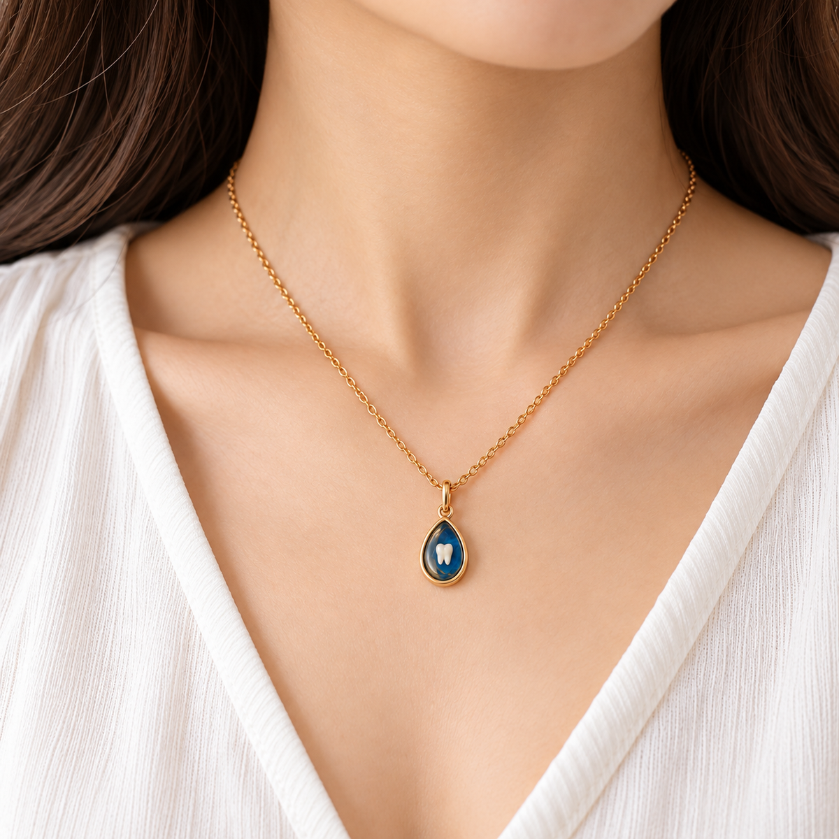 A close-up of a woman wearing a delicate gold chain necklace with a teardrop-shaped pendant featuring a deep blue enamel background and a white tooth motif in the center.